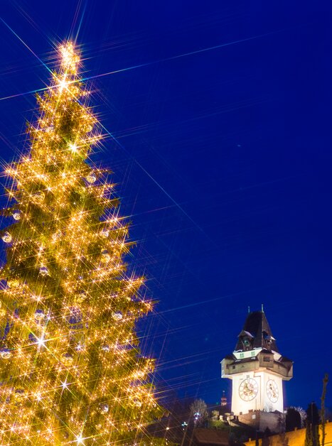 A beautifully illuminated Christmas tree with the Graz Clock Tower in the background, bright against the evening sky. | © Graz Tourismus - Harry Schiffer
