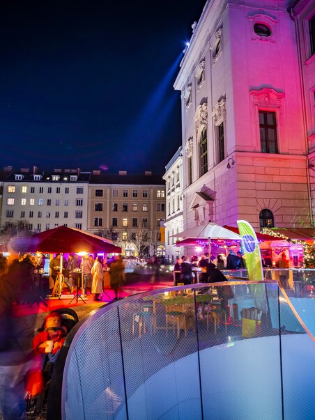 Verschiedene Stände und festliche Beleuchtung im Joanneumsviertel von Graz, umgeben von historischen Gebäuden und einer lebendigen Atmosphäre. | © Graz Tourismus - Werner Krug