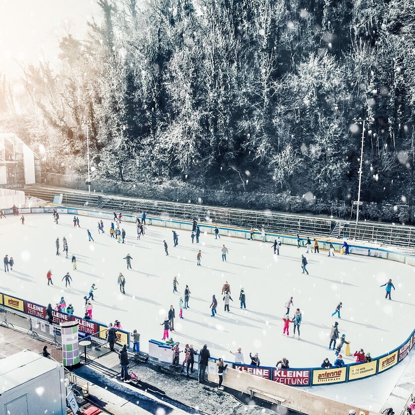 Menschen beim Eislaufen auf einem beleuchteten Eislaufplatz in Graz, umgeben von winterlicher Landschaft. | © Simon Möstl