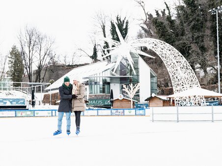 Ein Paar beim Eislaufen in der Winterwelt in Graz, mit Weihnachtsdekoration im Hintergrund. | © Graz Tourismus - Mias Photoart