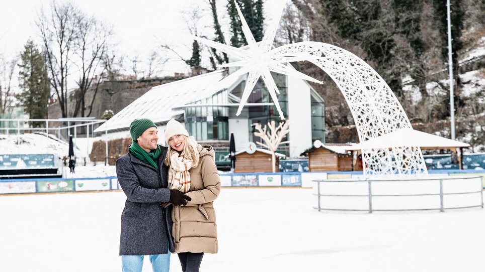 Couple on an ice rink in Graz, surrounded by a winter atmosphere and festive decorations. | © Graz Tourismus - Mias Photoart
