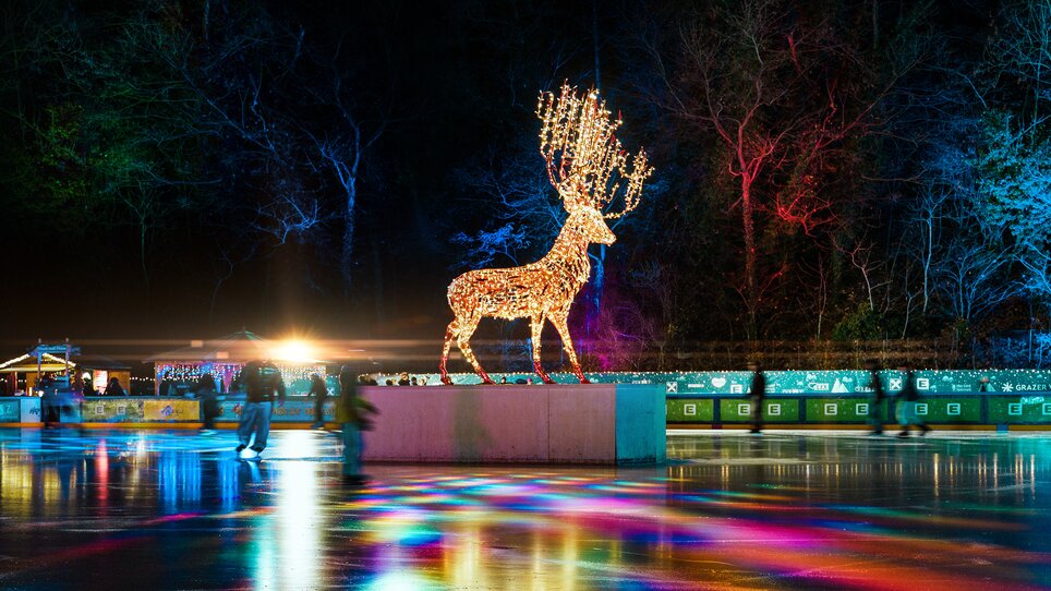 Illuminated reindeer at ice skating rink at night, surrounded by colorful lights and visitors. | © Leon Moik