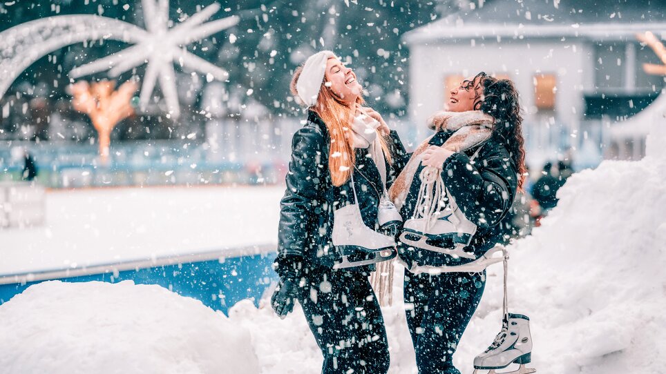 Two women laughing while holding ice skates in a winter wonderland. | © Stefan Leitner
