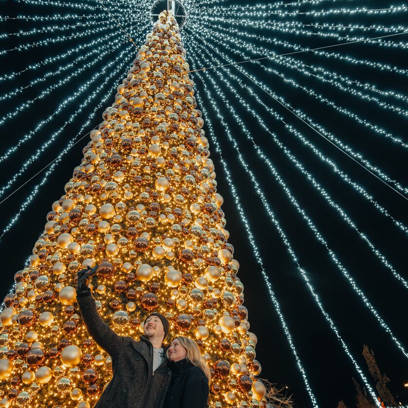 Advent in Graz I Winter Wonderlend am Mariahilferplatz | © Mias Photoart Ein beleuchteter Weihnachtsbaum mit Pärchen, die ein Selfie machen. | © Mias Photoart