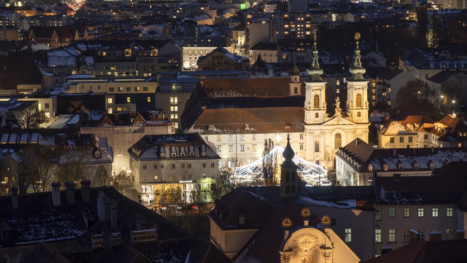 Città illuminata di Graz di notte con neve sui tetti e Mariahilferplatz in primo piano. | © Vincent Croce