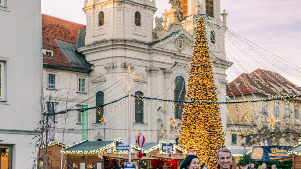 Una coppia passeggia sulla Mariahilferplatz a Graz, circondata da luci e un albero di Natale. | © Graz Tourismus - Mias Photoart