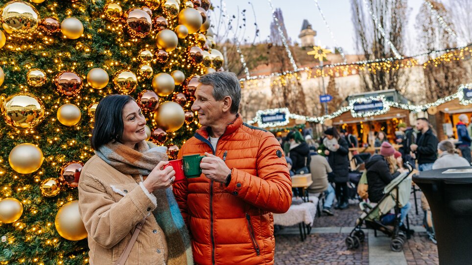 Coppia sorridente con tazze davanti all'albero di Natale e al mercato a Graz. | © Graz Tourismus - Mias Photoart
