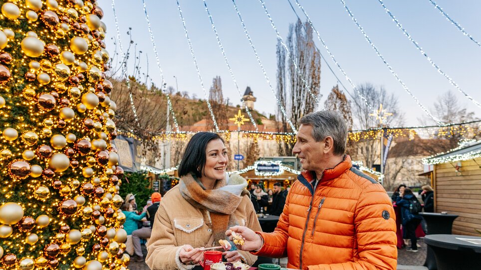 Una coppia si gode snack e bevande davanti a un grande albero di Natale in Mariahilferplatz a Graz. | © Graz Tourismus - Mias Photoart