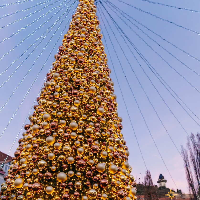 Ein großer Weihnachtsbaum mit goldenen Kugeln auf dem Mariahilferplatz in Graz, umgeben von einer festlichen Atmosphäre und dem Uhrturm im Hintergrund. | © Graz Tourismus - Mias Photoart