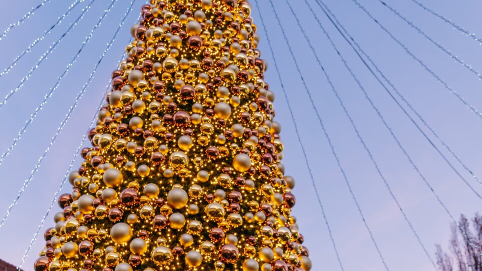 Un grande albero di Natale con palline dorate sulla Mariahilferplatz a Graz, circondato da un'atmosfera festosa e con la torre dell'orologio sullo sfondo. | © Graz Tourismus - Mias Photoart