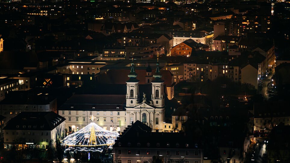 Mercato di Natale illuminato a Graz di notte, con la chiesa sullo sfondo. | © Graz Tourismus - Mias Photoart