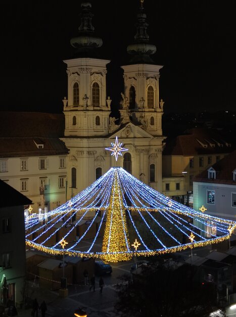 Illuminated Christmas market at Mariahilferplatz in Graz at night, surrounded by a church. | © Easy Conceptz GmbH
