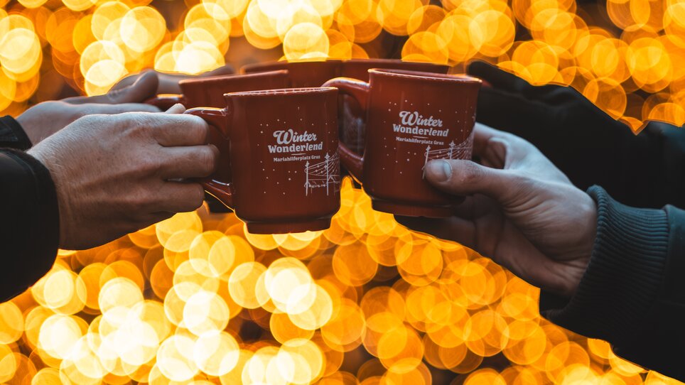 Four hands clink orange mugs, with glowing lights in the background. | © Easy Conceptz GmbH