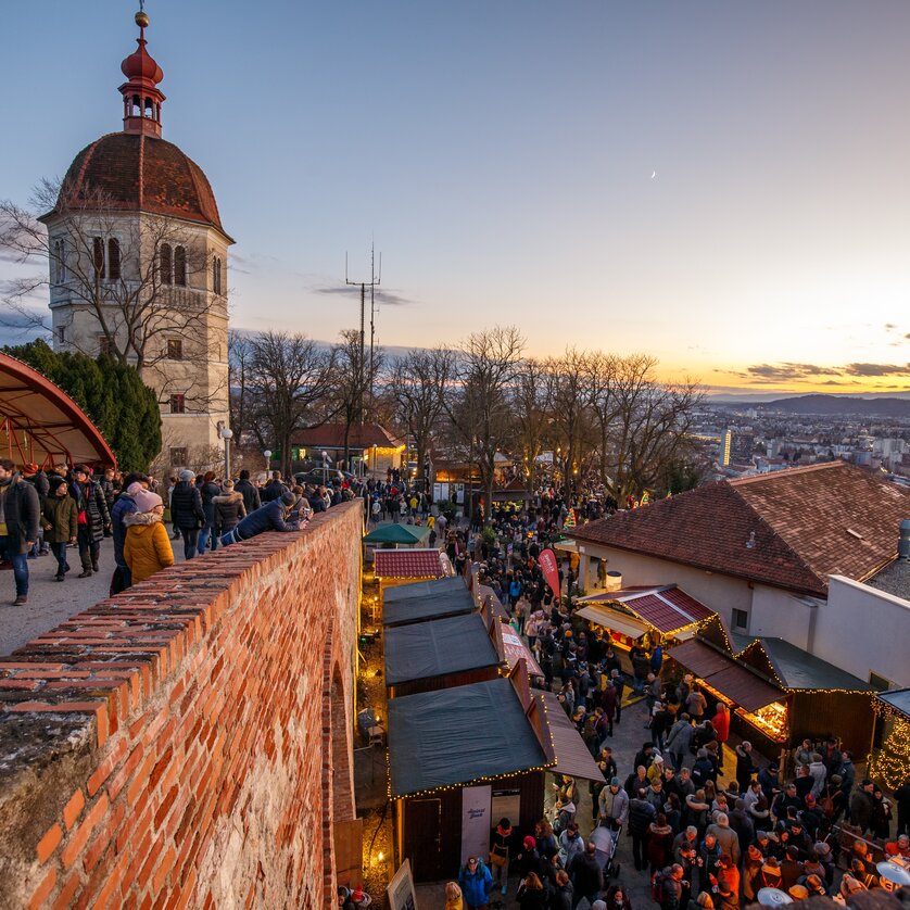 Vista del mercatino di Natale allo Schlossberg di Graz con molte persone e uno splendido tramonto sullo sfondo. | © ivents - Erwin Scheriau