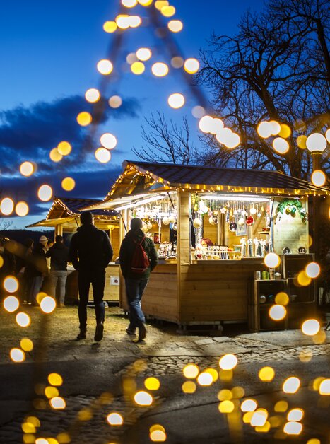 Weihnachtsmarkt mit bunten Lichtern und Ständen, unter blauem Himmel. | © ivents - Erwin Scheriau
