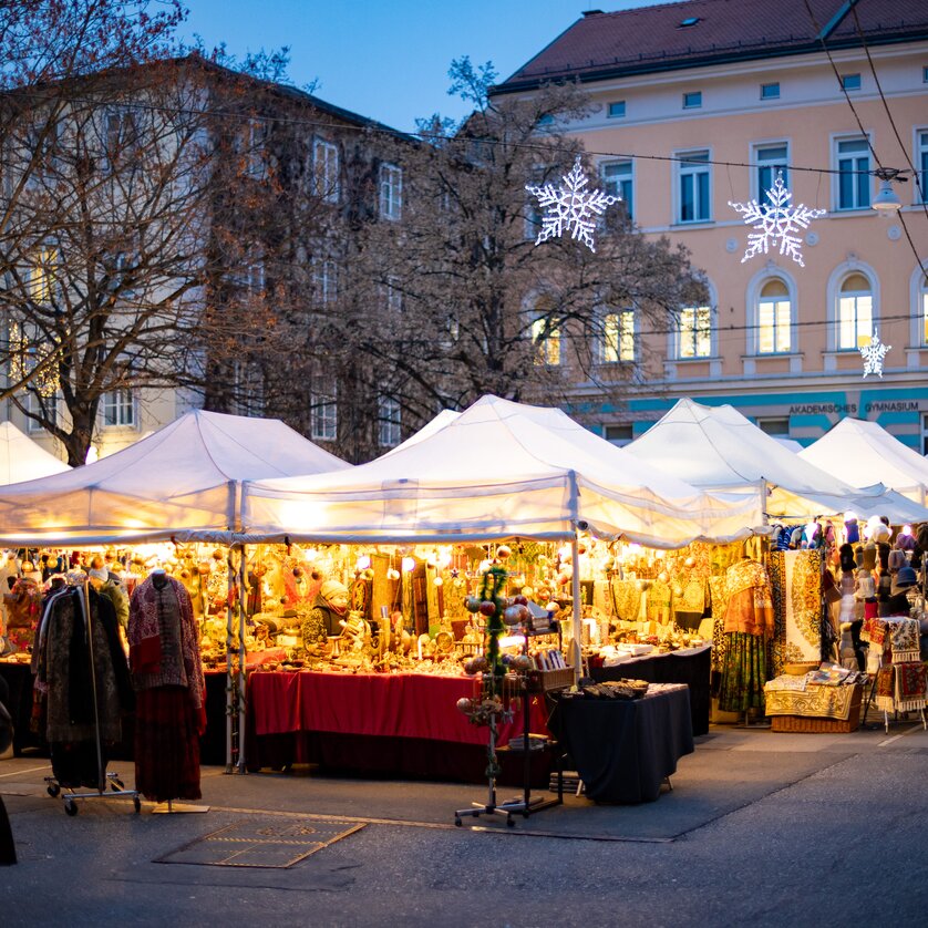 Una scena festiva del mercatino di Natale con stand illuminati e gente a Graz. | © Graz Tourismus - Harry Schiffer