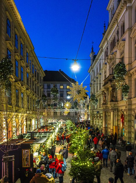 Illuminated Advent stalls and Christmas market in Schmiedgasse, Graz, festive evening with many visitors. | © Rene Walter