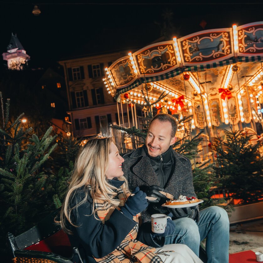 Advent in Graz I Christmas on Karmeliterplatz | © Mias Photoart A couple enjoys Christmas treats in front of a beautifully lit carousel in Graz. | © Mias Photoart