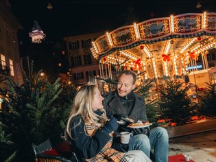Ein Paar genießt Weihnachtsleckereien vor einem festlich beleuchteten Karussell in Graz. | © Mias Photoart