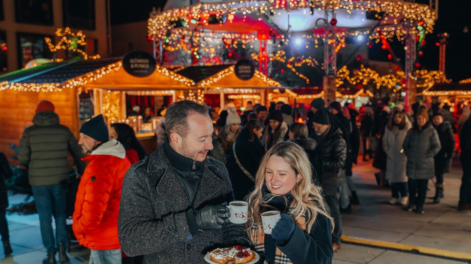 Ein Paar genießt Glühwein und Gebäck auf einem festlich beleuchteten Weihnachtsmarkt. | © Mias Photoart