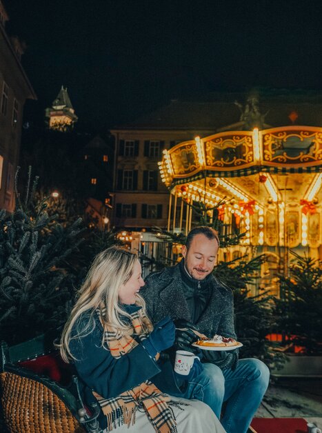 A couple enjoys the Christmas atmosphere at Karmeliterplatz in Graz with a carousel in the background. | © Mias Photoart