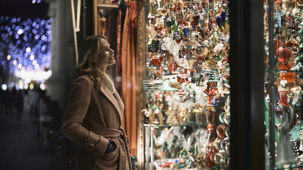 Eine Frau steht vor einem Schaufenster voller weihnachtlicher Ornamente und schaut bewundernd auf die bunten Dekorationen. | © Robert Maybach