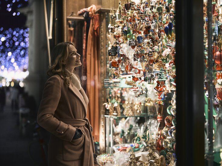 Eine Frau steht vor einem Schaufenster voller weihnachtlicher Ornamente und schaut bewundernd auf die bunten Dekorationen. | © Robert Maybach