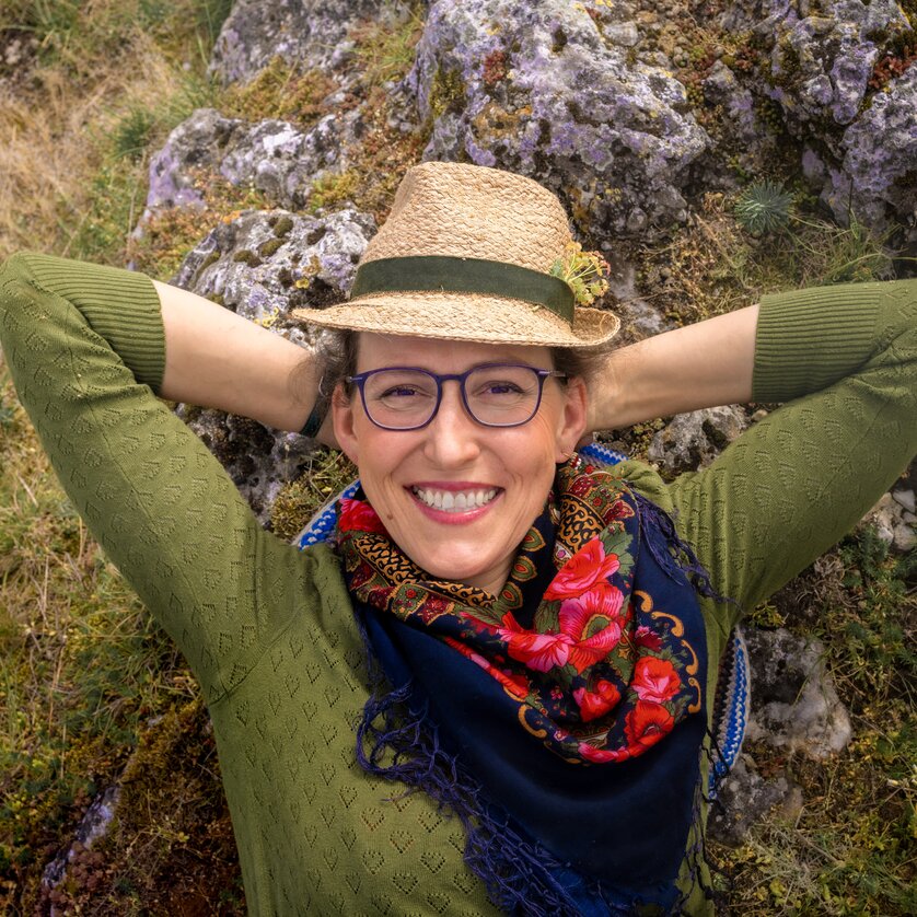Frau mit Hut und Schal lächelt entspannt in der Natur.