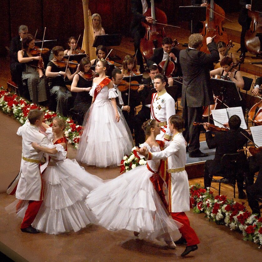 Tänzer in eleganten Kostümen führen eine Darbietung vor einem Orchester auf.