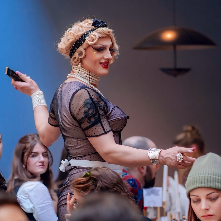 Ein Drag-Künstler posiert in einem Restaurant, umgeben von Gästen. | © Bernhard Schinder
