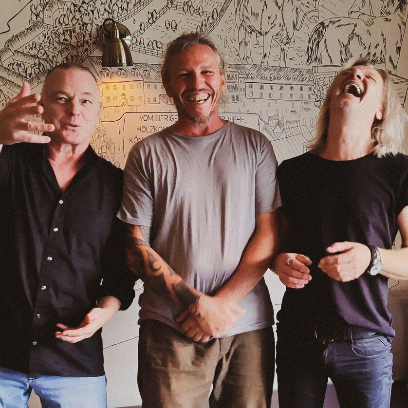 Drei Männer lachen und posieren vor einer Wand mit Zeichnungen. | © Andre Schwarzl