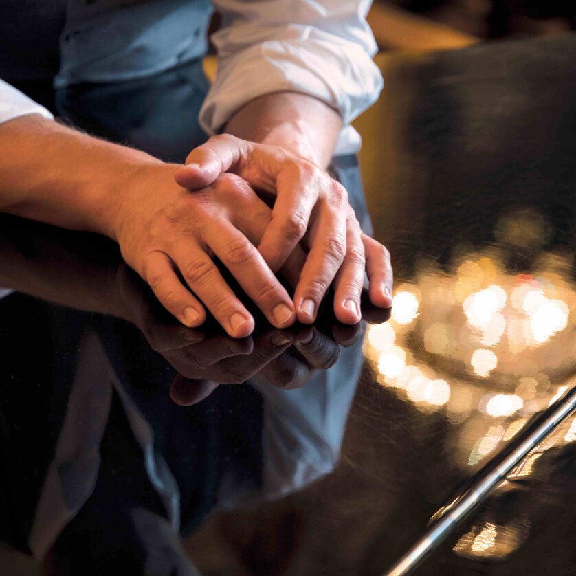 Die Hände einer Person ruhen auf einem glänzenden Klavier, am Klavier spiegelt sich eine Kronleuchter-Beleuchtung. | © Pilo Pichler