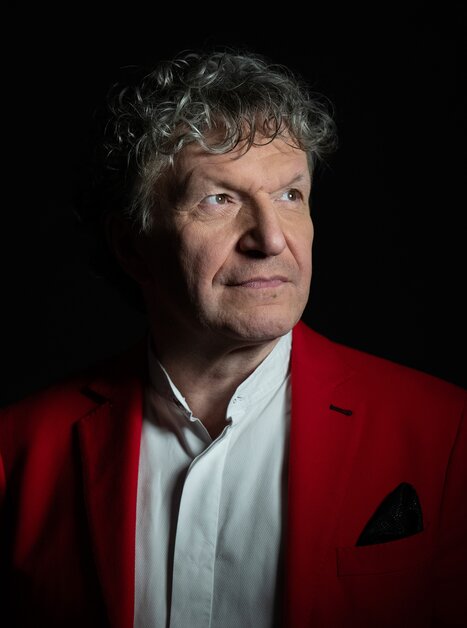 Ein Mann mit lockigem Haar in einem roten Blazer, frontal beleuchtet, vor dunklem Hintergrund. | © Christian_Jungwirth_verk