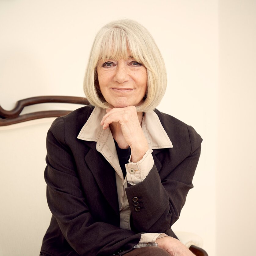 Erika Pluhar sits on a white armchair in an elegant suit, smiling. | © Lukas Beck