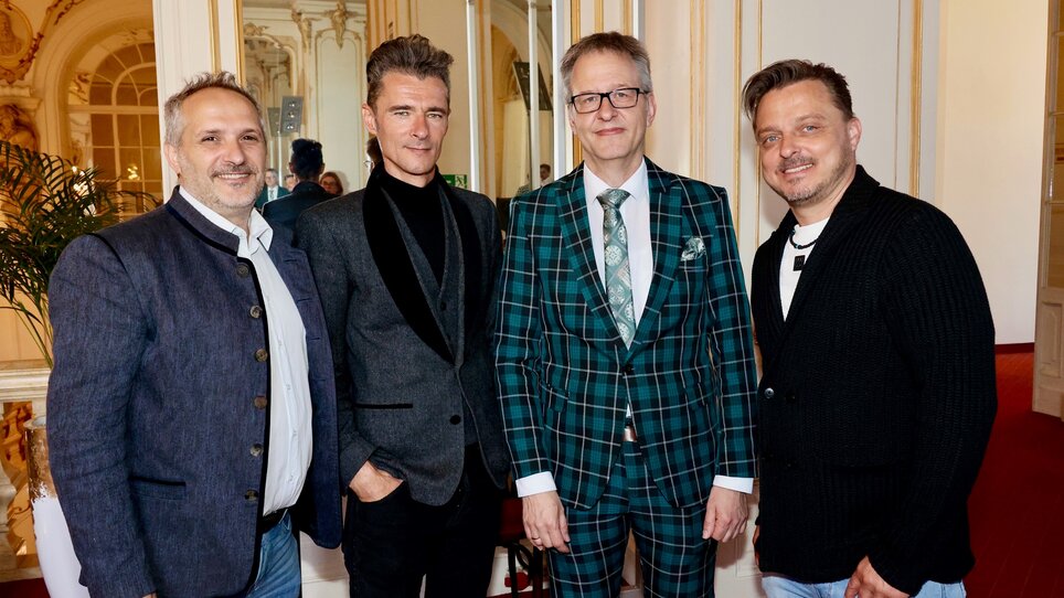 Four men stand in a magnificent room with high ceilings, mirrors and golden decorations. They are all wearing eye-catching outfits: from traditional Styrian-inspired jackets and elegant jackets to a chequered suit and tie. They look friendly into the camera. | © Ivents Kulturagentur