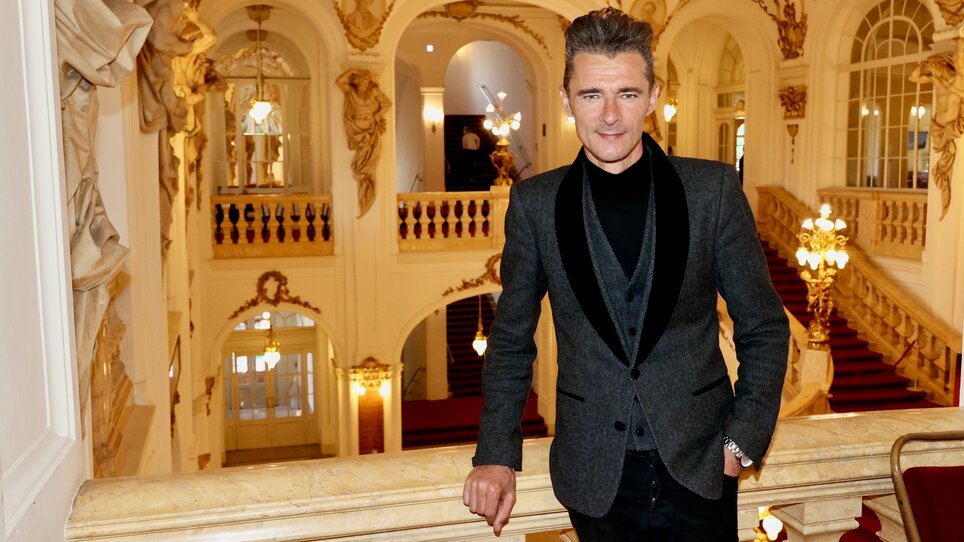 Herbert Pixner leans against the balcony railing of the Graz Opera House, behind him you can see the staircase with the red carpet. | © Ivents Kulturagentur