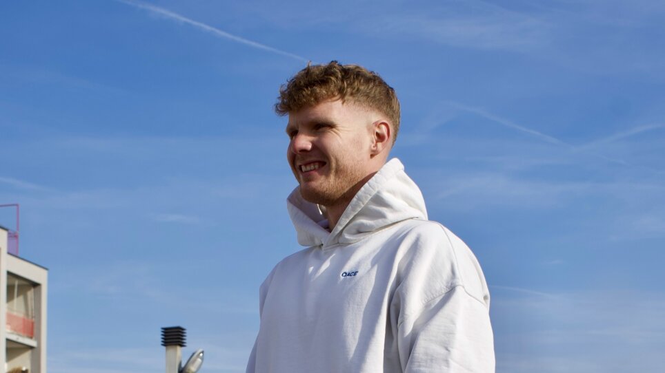 Ein junger Mann in legerer Sportkleidung steht auf einer Dachterrasse und lächelt  | © Alex Eibinger