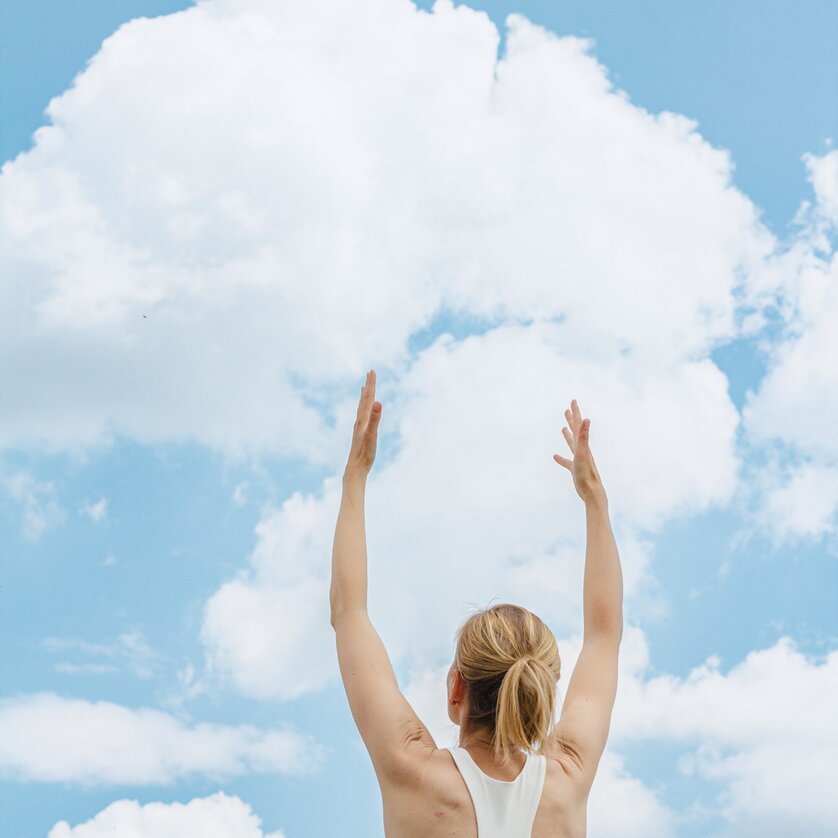 Una donna bionda in abbigliamento sportivo allunga entrambe le braccia verso l'alto, alle sue spalle si intravede un cielo blu con nuvole | © Augartenhotel x einsnullneun