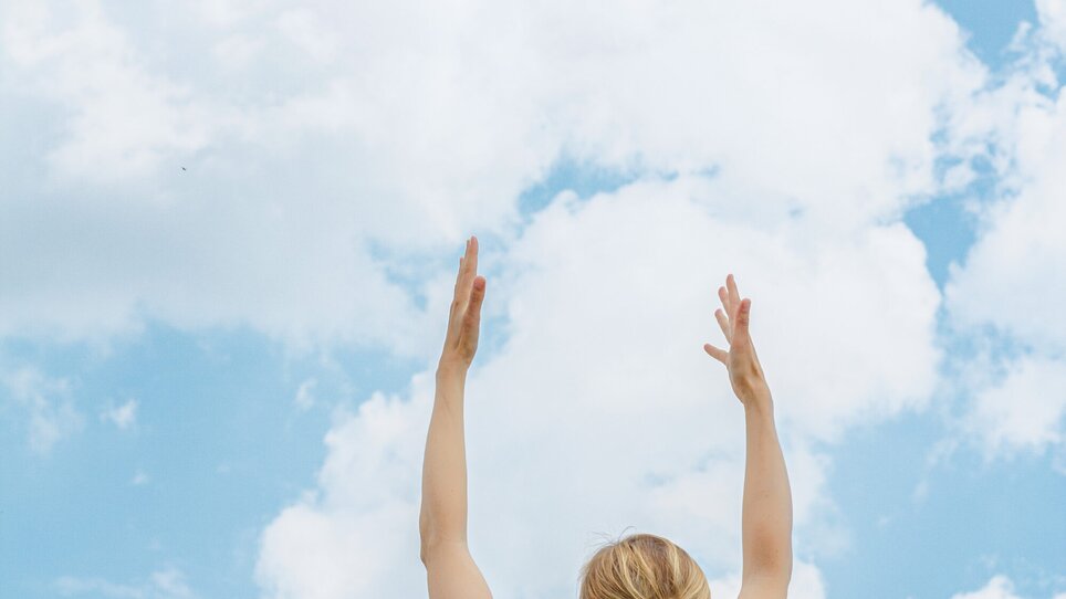 A blonde woman in sportswear stretches both arms upwards, a blue sky with clouds can be seen behind her | © Augartenhotel x einsnullneun
