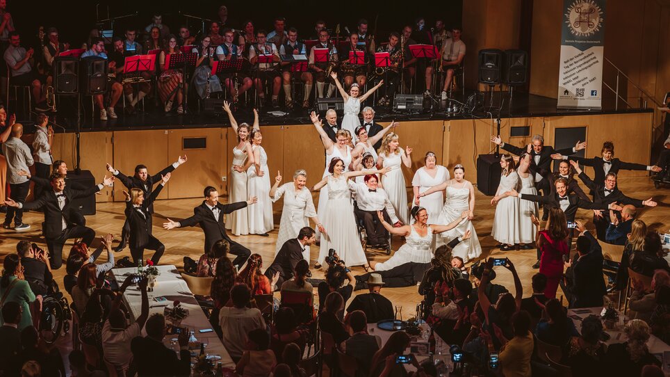 Tänzer und Tänzerinnen in schwarzen Anzügen und weißen Kleidern posieren nach dem Abschluss ihrer Darbietung vor einer Bühne | © Mario Sudy