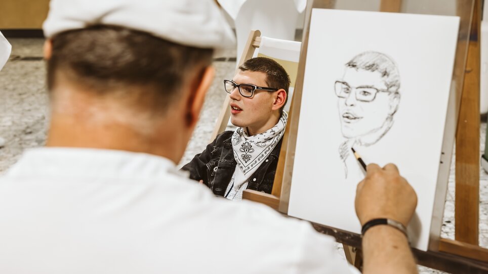 Über die Schulter des Porträtzeichners hinweg sieht man einen jungen Mann und sein Porträt, das gerade angefertigt wird | © Mario Sudy