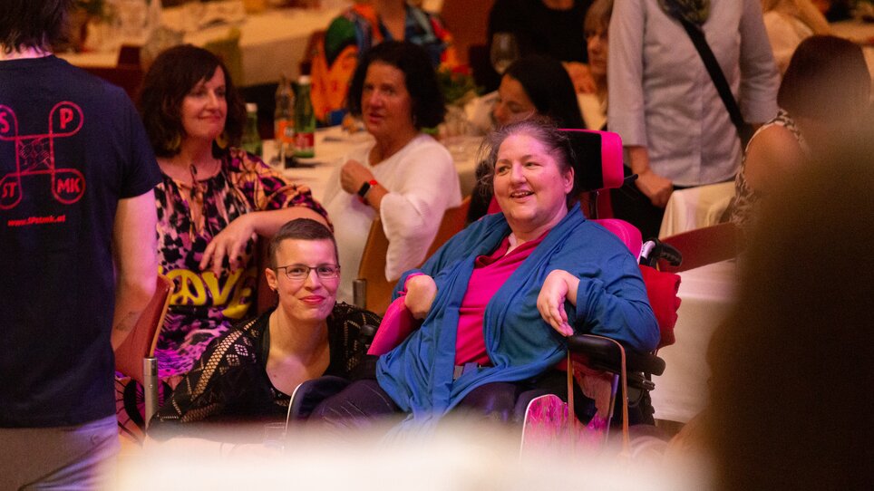 Zwei Frauen beobachten das Bühnenprogramm. Eine von ihnen sitzt im Rollstuhl, die andere kniet neben ihr am Boden. | © Péter Grátz