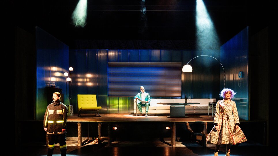 Drei Darsteller in auffälligen Kostümen auf einer Theaterbühne.