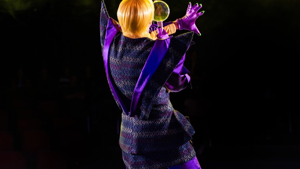 Eine Person in einem auffälligen, lila Outfit steht auf der Bühne und hält ein Mikrofon.
