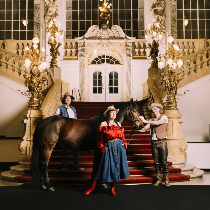 Drei Menschen in Cowboykleidung mit Pferd auf rotem Teppich in prunkvollem Opernfoyer | © Oper Graz - Ingo Pertramer