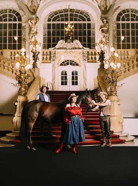 Drei Menschen in Cowboykleidung mit Pferd auf rotem Teppich in prunkvollem Opernfoyer | © Oper Graz - Ingo Pertramer