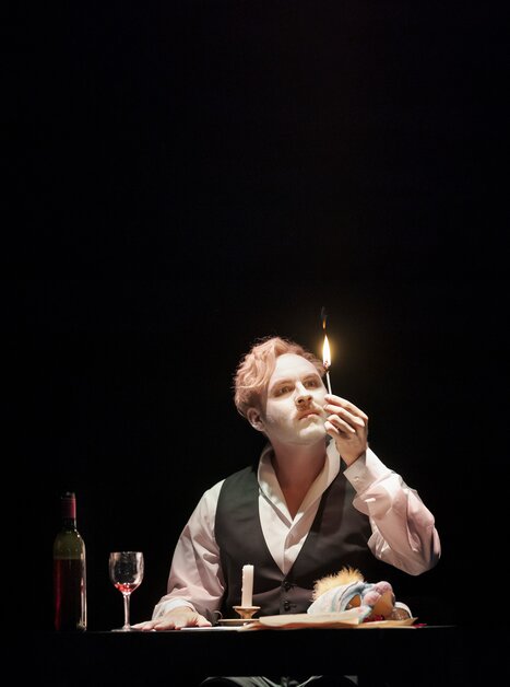 An actor with a painted face holding a lit candle during a performance. | © Oper Graz - Werner-Kmetitsch