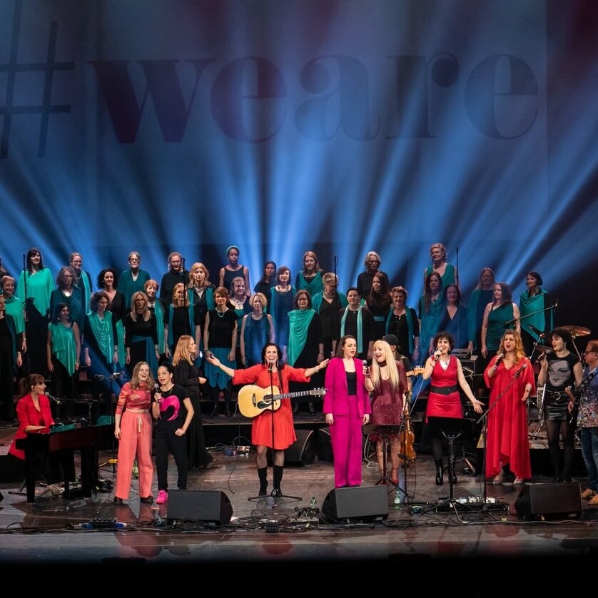 weare | © Iva Lulic Große Gruppe von Sängerinnen und Musiker:innen bei Konzert mit Bühnenlicht und #weare-Backdrop | © Iva Lulic