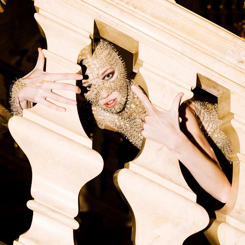 La Divina Comedia Kirsty-Clarke | © Oper Graz - Ingo Pertramer A person in a gold mask poses behind a balustrade-like structure. | © Oper Graz - Ingo Pertramer