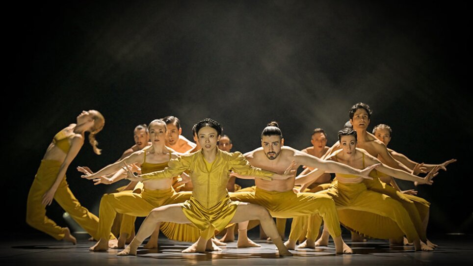 A group of dancers in yellow costumes performing synchronized movements on stage. | © Andreas J. Etter - Oper Graz